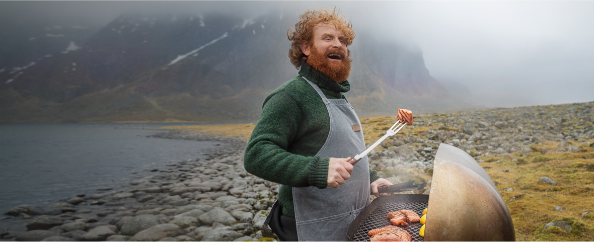 Un homme barbu cuisine au barbecue au bord d'un lac de montagne brumeux.