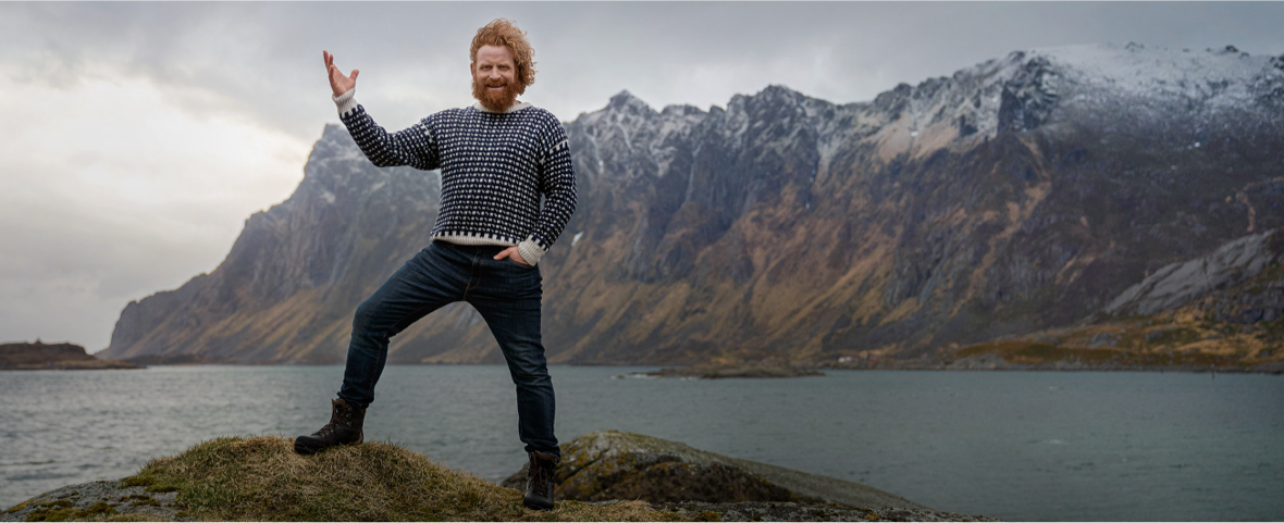 Homme roux au pull norvégien levant la main avec des montagnes enneigées en arrière-plan.