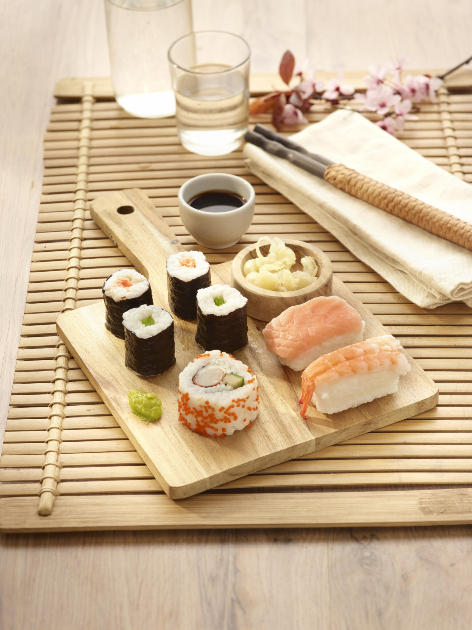 Assortiment de makis, nigiris, wasabi et gingembre sur un plateau en bois, avec des baguettes et une branche de cerisier en fleur.