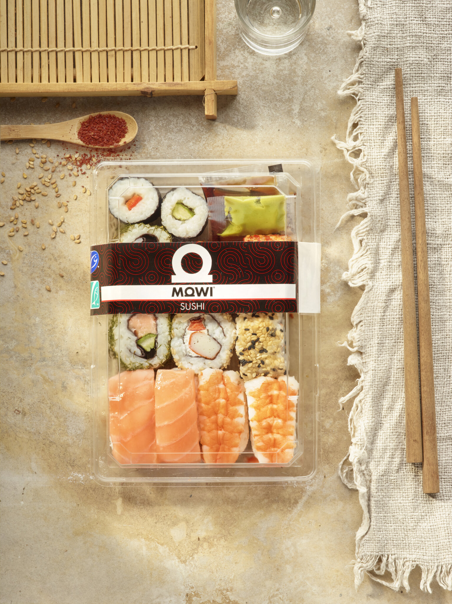 Assortiment de sushis dans un emballage transparent avec baguettes et condiments sur fond texturé.